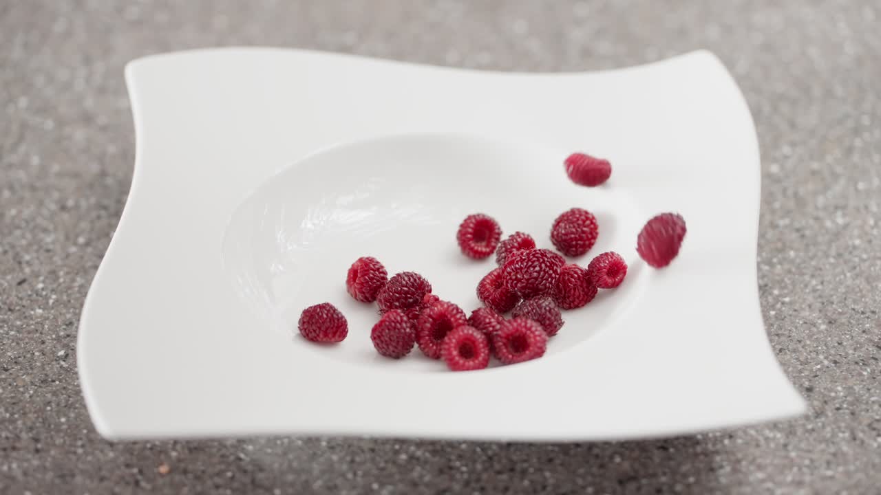 Close-up of organic fresh raspberries dropping into a stylish white plate, capturing the dynamic motion for concepts of healthy desserts and frozen fruit ingredients - slow motion