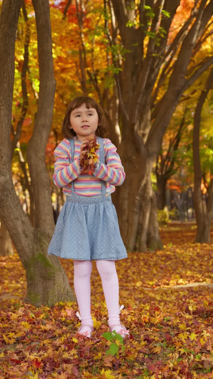 Vertical - Joyful little girl with a happy smile picks up colorful autumn maple leaves from the ground and playfully throws them into the air in a vibrant park