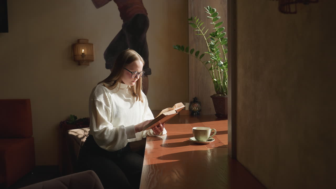 Real estate mogul in white sweater reading novel thoughtfully near sunlit window with potted plant and coffee cup on wooden table, immersed in quiet ambiance and warm natural light indoors