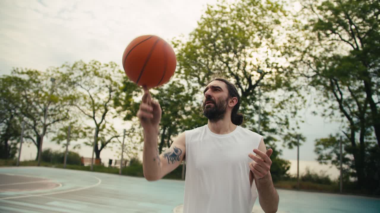 un hombre de mediana edad con una camiseta blanca hábilmente gira una pelota de baloncesto naranja en su dedo índice en un campo deportivo en verano