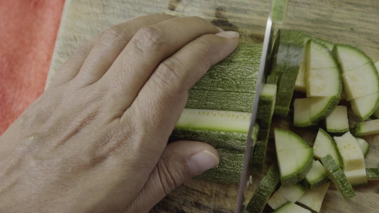Primo piano di una mano che taglia zucchine fresche a fette su un tagliere di legno, una semplice scena di cucina che evidenzia la preparazione degli ingredienti e un processo di cottura sano.