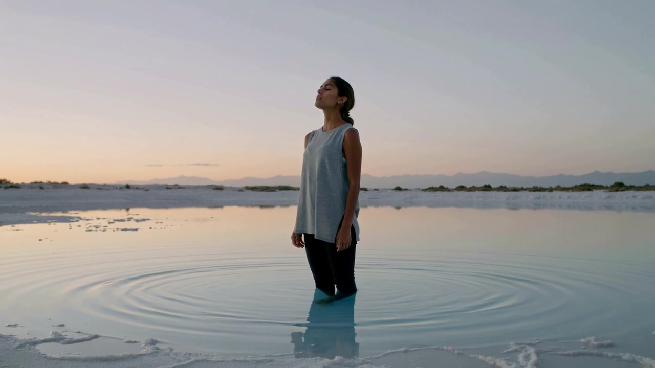 Con il mento inclinato, una donna osserva l'alba in piedi fino alle ginocchia in una piscina increspata, indossando una canotta e pantaloni scuri.