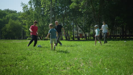 grandchildren are playing with a soccer ball in a grassy field as their grandfather joins in, trying to get the ball