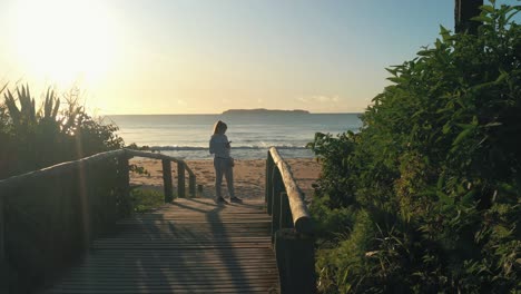 beach sunrise golden hour. woman stands on wooden deck using smartphone with running suit and sneakers. dolly shot