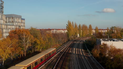 Personenzug-Mit-Graffiti-Vorbei-An-Bäumen-In-Der-Großstadt-Berlin,-Deutschland-Bei-Sonnenuntergang-Zur-Goldenen-Stunde,-Stadtbild-Mit-öffentlichen-Verkehrsmitteln-Neben-Bürogebäude,-Luftwagen-Herein