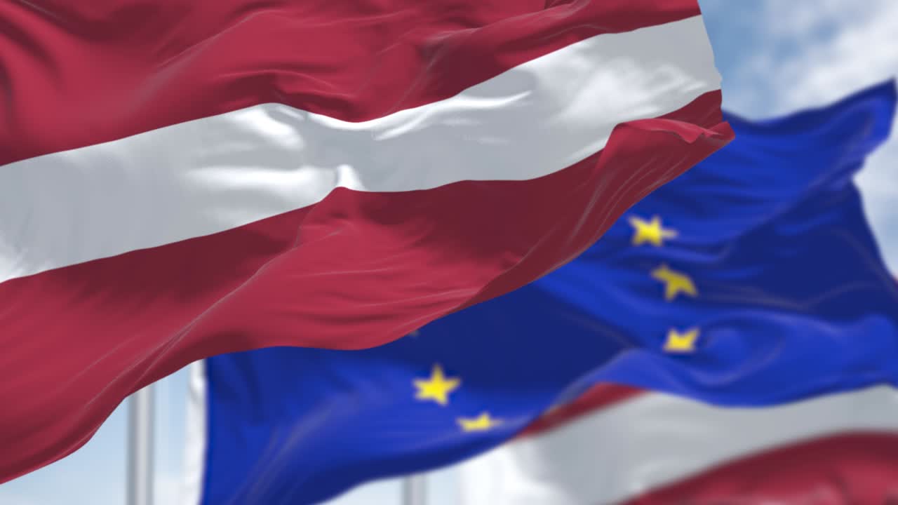 라트비아의 국기가 바람에 흔들리고, 배경에는 유럽연합의 발이 흐릿하다.