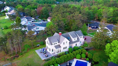 Beautiful aerial drone footage of a cozy cottage-style suburban house with a big back porch, comfy patio seating, and a green lawn, all enclosed by tall, mature, leafy trees in a peaceful neighborhood