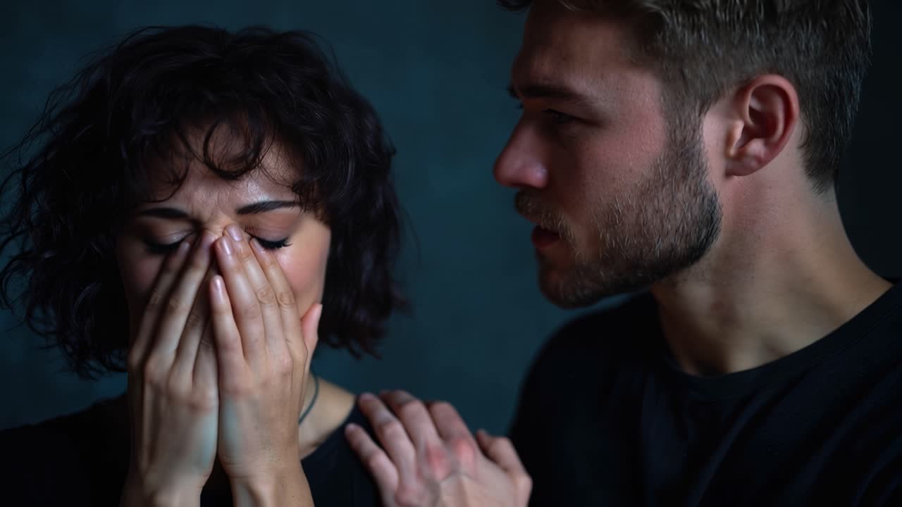 Una donna si copre la bocca, gli occhi spalancati per lo shock. L'uomo accanto a lei guarda in basso, con una mano sulla sua spalla. L'oscurità li circonda, enfatizzando il loro momento emotivo