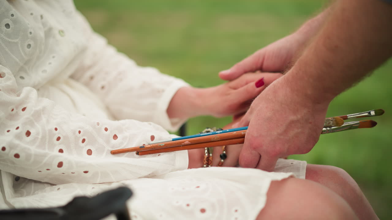 예술가의 손이 여성의 손을 부드럽게 조정하는 클로즈업, 그녀의 반지와 구슬로 된 팔를 강조합니다. 이 장면은 신중한 관심과 예술의 친밀한 순간을 포착합니다.