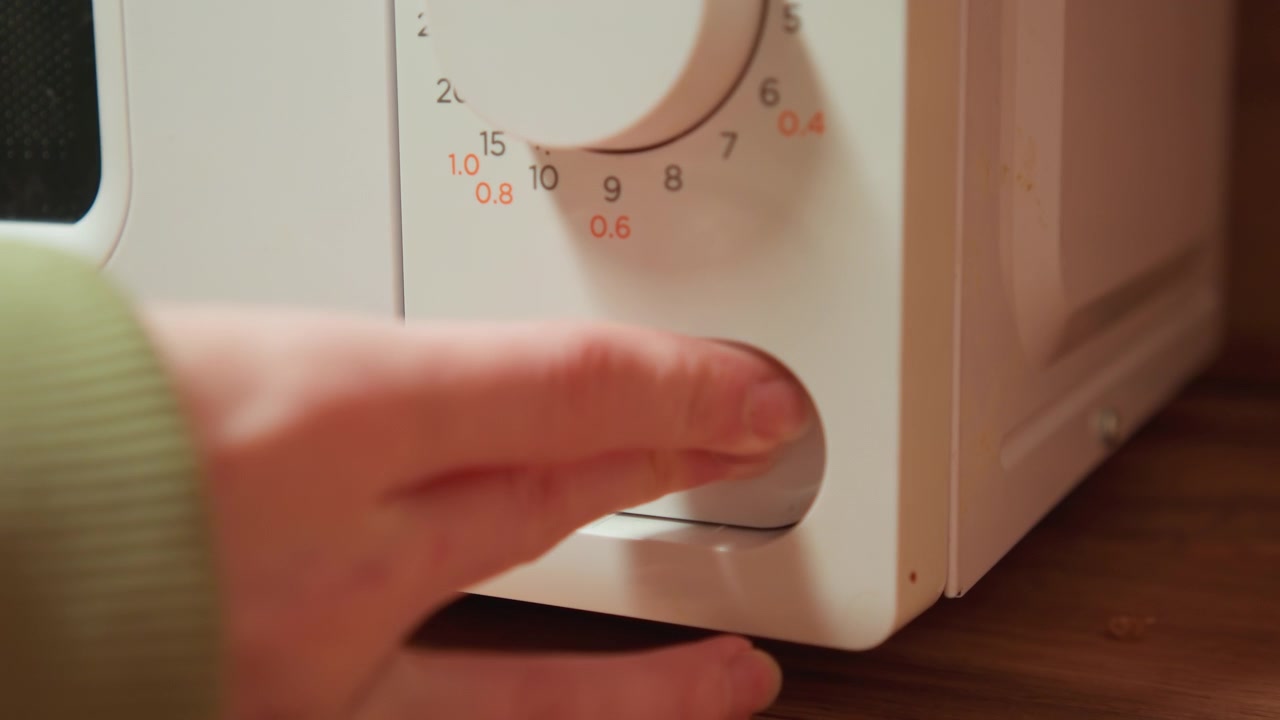 Primo piano di una mano che gira la manopola del timer del microonde mentre le punte delle dita premono il pannello di controllo inferiore su un rivestimento in plastica bianca posizionato sopra un piano di lavoro in legno, mostrando una semplice regolazione della cucina in un ambiente interno caldo.