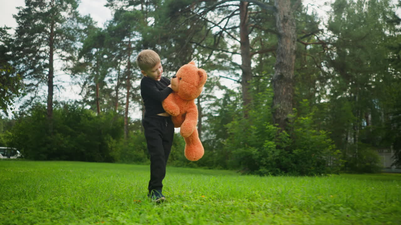Young child dressed in black standing on green grass holding large orange teddy bear, looking down thoughtfully, with parked bus and forest trees in background