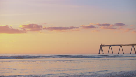 Ein-Dramatischer-Sonnenuntergang-Am-Orangefarbenen-Strand-Mit-Leichten-Wolken-Und-Einem-Steg-In-Der-Ferne