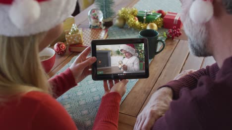 Smiling-caucasian-couple-with-santa-hats-using-tablet-for-christmas-video-call-with-boy-on-screen