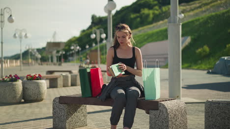 lady seated on outdoor bench with shopping bags on both sides, retrieves a book from mint green bag, opens it, and goes through its pages, background includes lamp poles, greenery