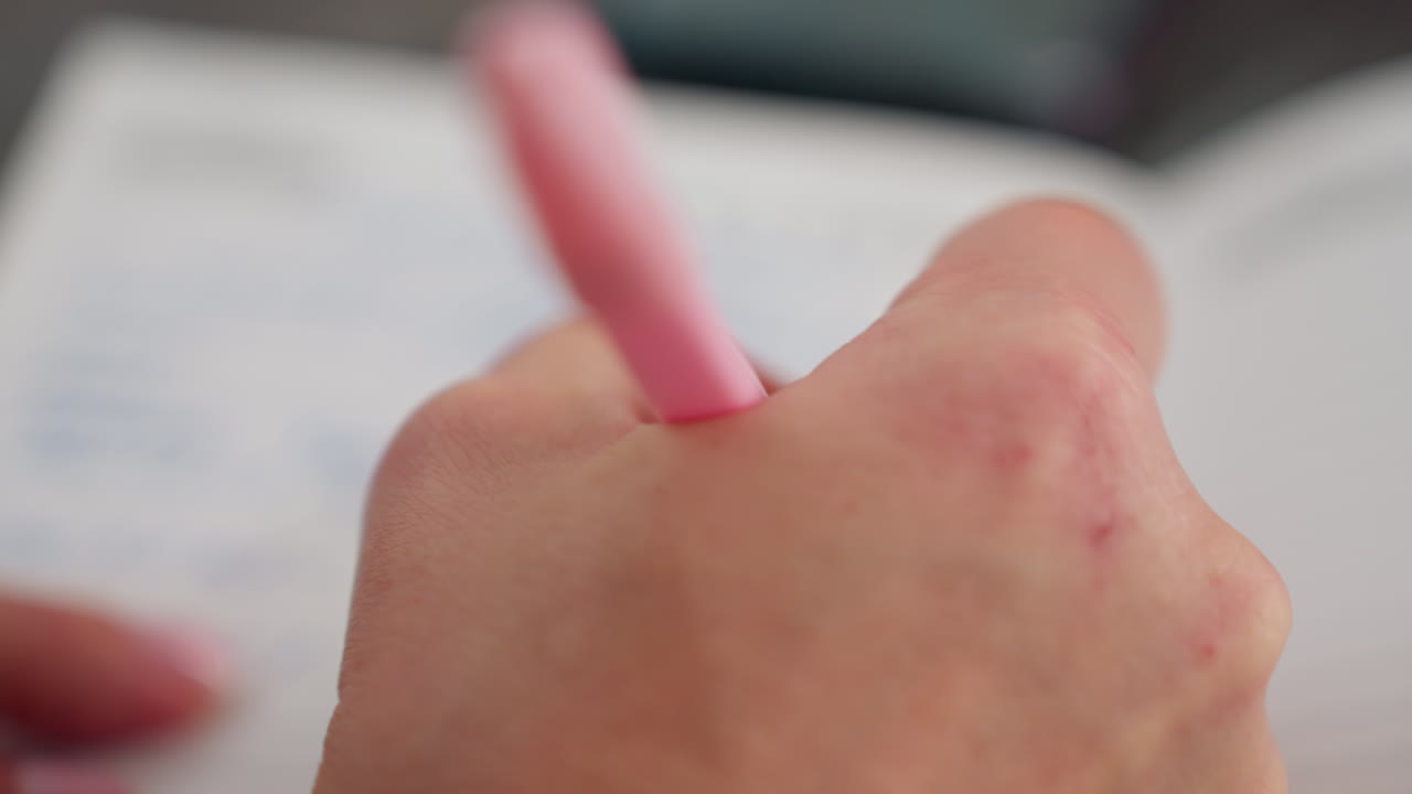 Close up hand view of white person writing with pink pen on paper with soft blur surroundings showing concentration and gentle motion, with natural lighting in background