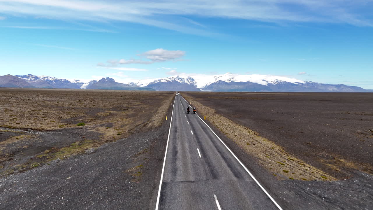 Dronebillede i fugleperspektiv af to cyklister, der cykler på en øde vej nær Vatnajökull-gletsjeren i Island, og krydser en enorm vulkansk slette med sneklædte bjerge under en klar, blå himmel