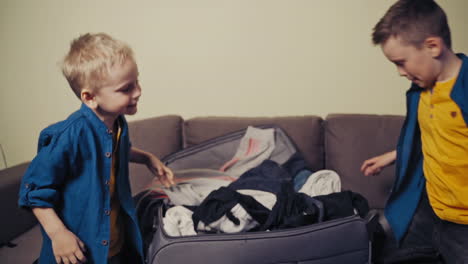 happy brothers packing a big suitcase with clothes at home. two cute boys in blue shirts putting many clothes into the travelling bag while sitting on the sofa in the room. slow motion.