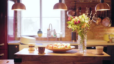 Hermosa-Cocina-Soleada-Con-Flores-Y-Una-Saludable-Canasta-De-Frutas-Para-El-Desayuno,-Llena-De-Plátanos,-Naranjas,-Fresas-Y-Uvas