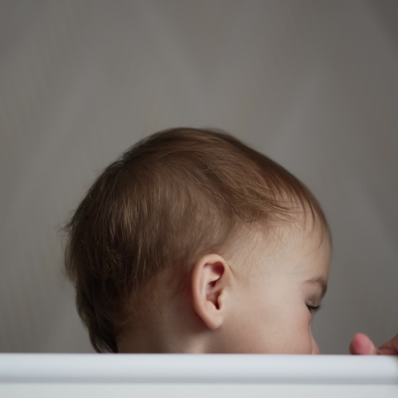 Cute adorable kid wearing grey shirt stands in cot holding by the handrail. Nice child sits in his crib hiding behind the bed border