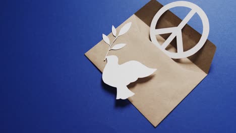 Close-up-of-white-dove-with-leaf,-white-peace-sign,-envelope-and-copy-space-on-blue-background