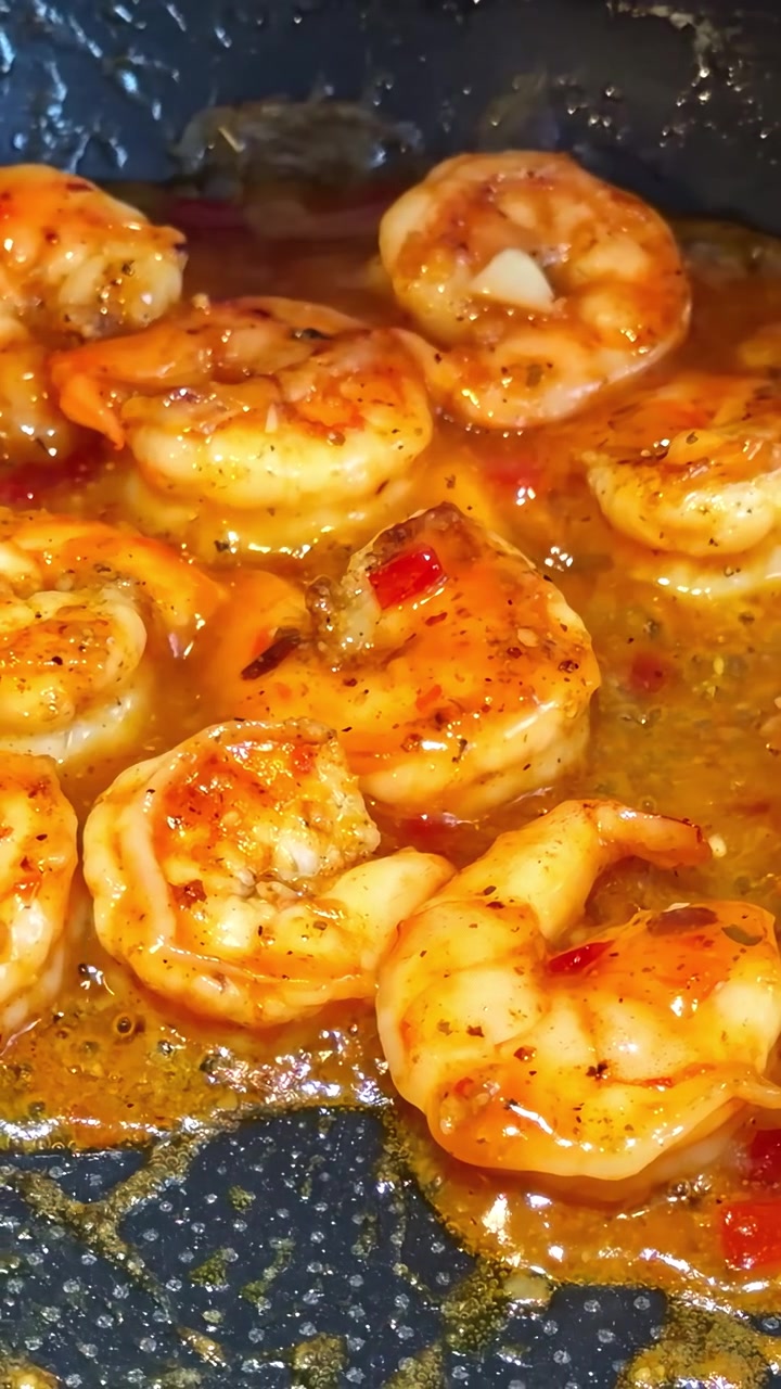 Gamberi che sobbollono in una salsa saporita e piccante mentre cuociono in una padella nera, mostrando vivaci tonalità arancioni e condimenti luccicanti. Video verticale