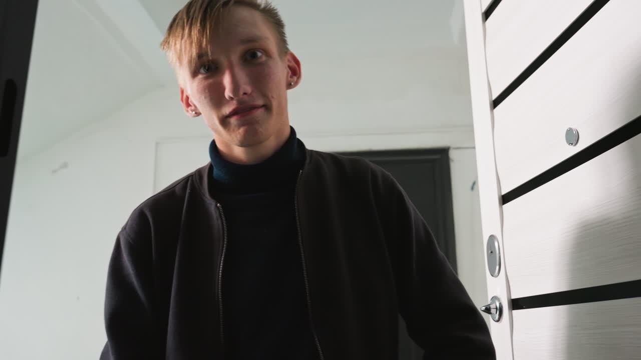 Jovem caucasiano segurando um pacote na porta do apartamento, sorrindo ao entrar na entrada mal iluminada, porta moderna com maçaneta de metal, verificando o rótulo do endereço, preparando a entrega sem contato, jaqueta casual