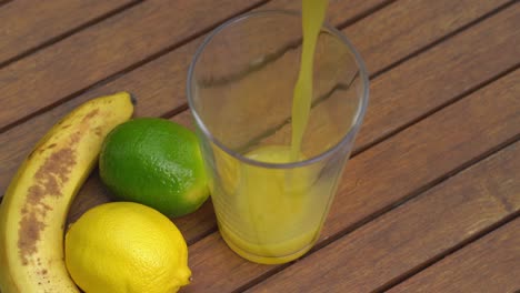 Pouring-glass-of-orange-juice-on-garden-table-with-colorful-Banana,-Lemon-and-Lime-fruit