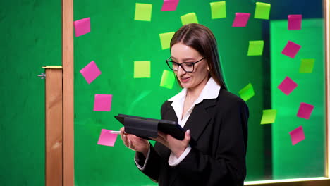 beautiful smiling lady wearing glasses, black jacket and white shirt stands near the glass board. woman talks to someone aside and then looks at her tablet.