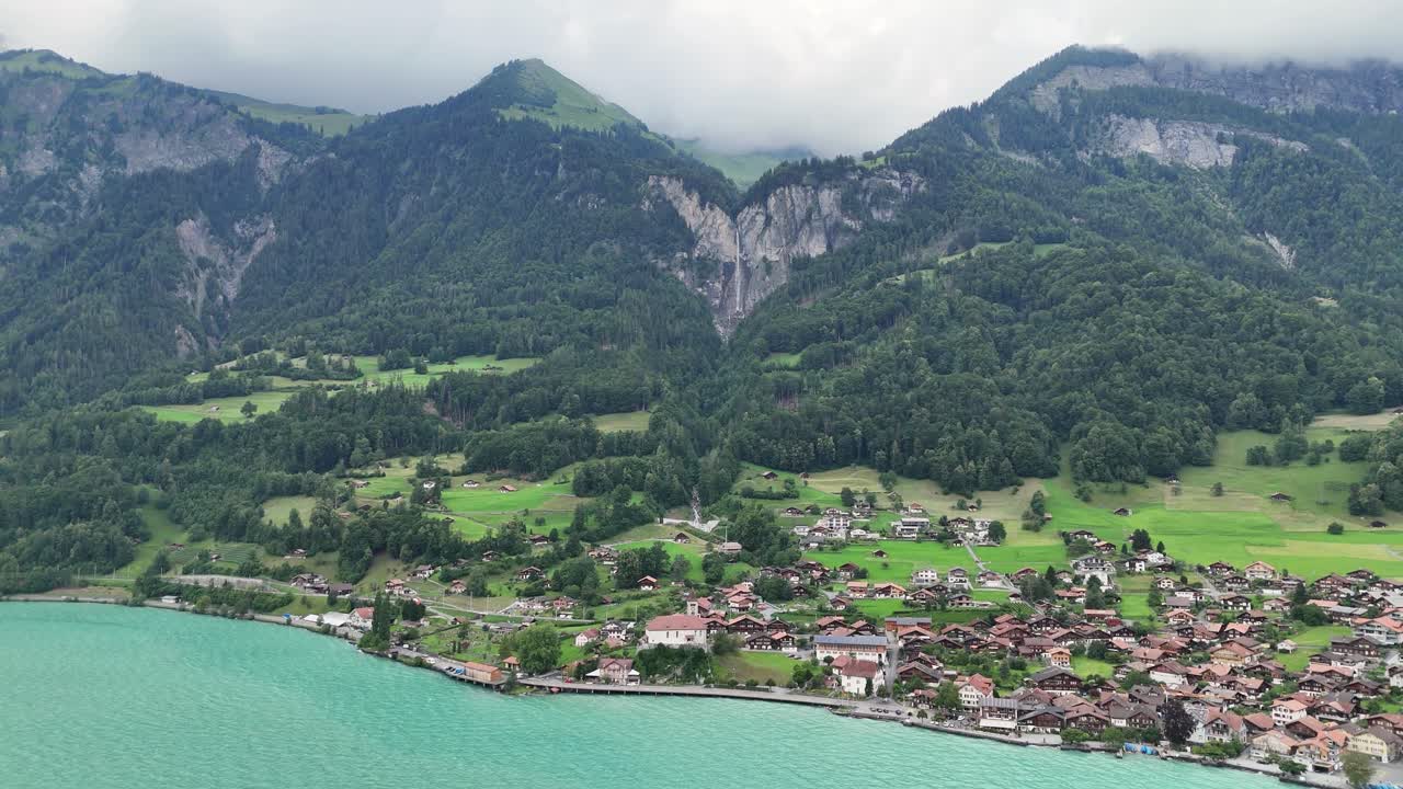 ドローンが、ターコイズブルーのブリエンツ湖に面したブリーンツ村上空を降下。スイスアルプスを背景に滝が流れ、曇り空の下、豊かな緑の山々が広がる。