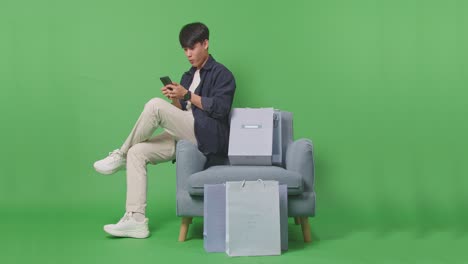 쇼핑 가방 을 들고 있는 아시아인 쇼핑맨 이 녹색 스크린 앞 에 소파 에 앉아 휴대 전화 로 온라인 쇼핑 을 즐긴다