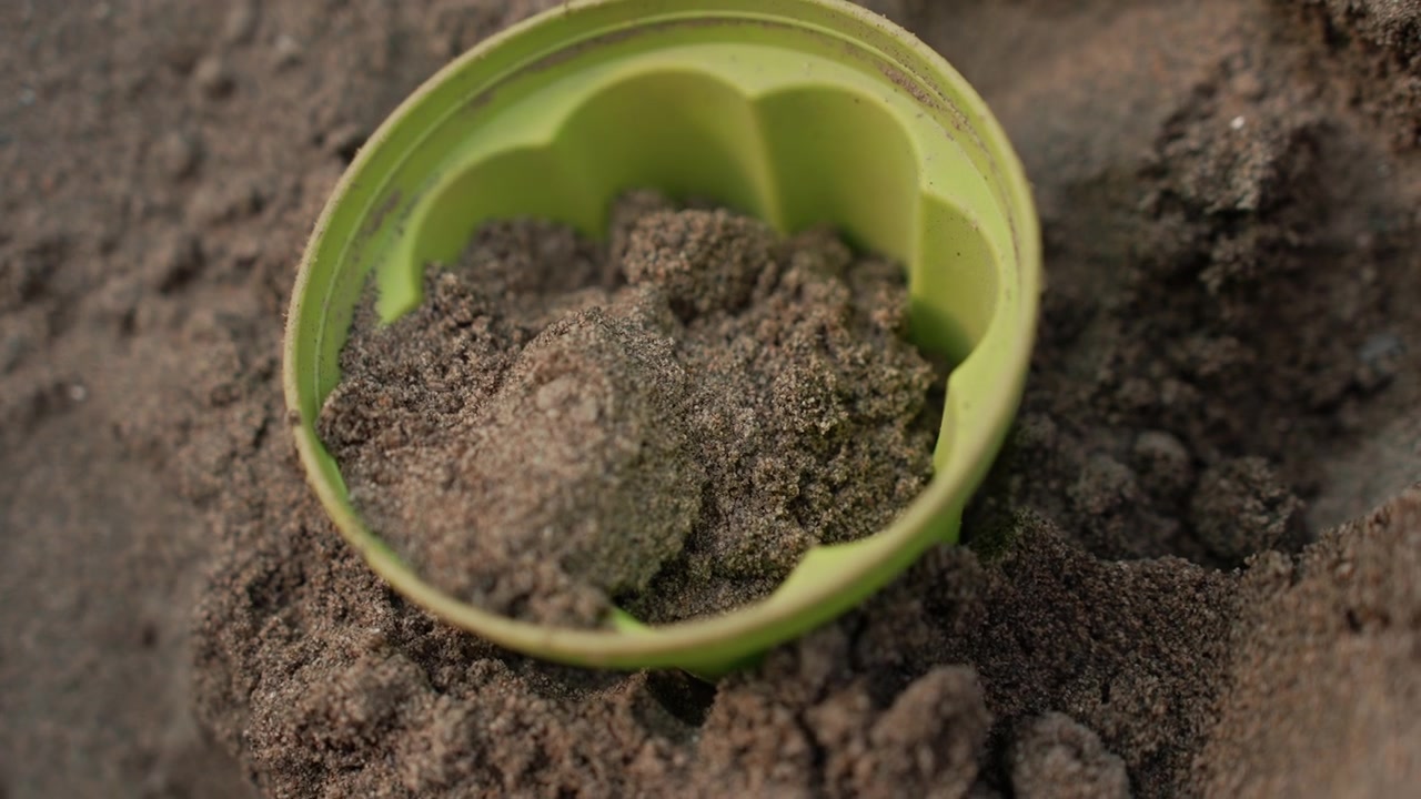 Small green plastic container is repeatedly filled with dark soil or sand as hand places clumps into it. background remains consistently out of focus emphasizing texture and detail