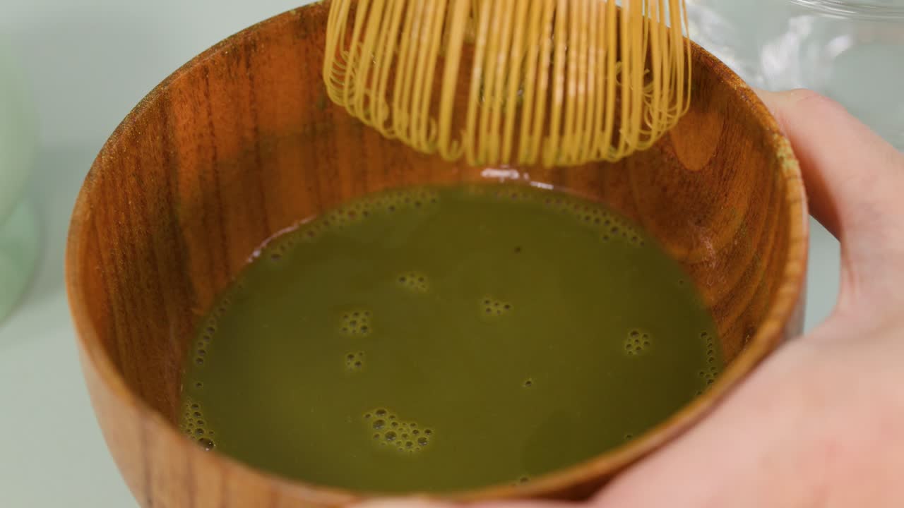Hand uses bamboo whisk to froth matcha green tea in wooden bowl, bright soft lighting
