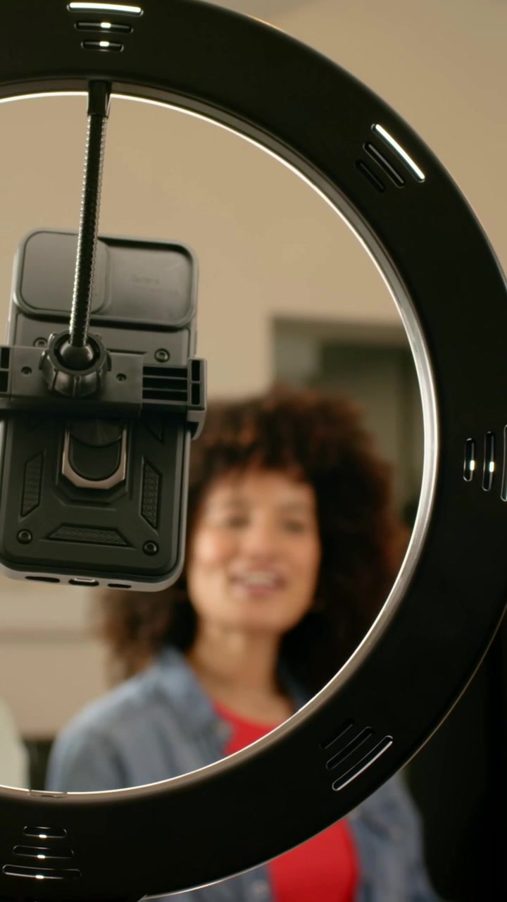 Video verticale: Donna con un top rosso che controlla il telefono sotto una ring light mentre la registrazione inizia in studio.