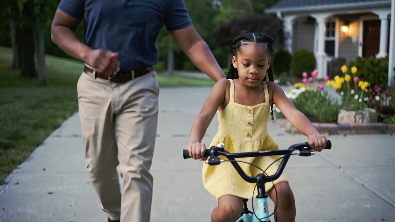 Learning girl yellow dress pedaling turquoise bike down driveway, father entering from left helping