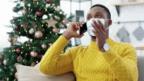 Cierra-El-Retrato-De-Un-Tipo-Alegre-Con-Máscara-Médica-Sentado-En-Un-Sofá-En-Una-Habitación-Decorada-En-Casa-Cerca-Del-árbol-De-Navidad-Y-Charlando-En-Un-Smartphone