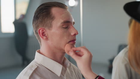 makeup artist preparing model face in minimalist studio using sponge and liquid foundation for even coverage while mirror captures soft natural lighting and room background