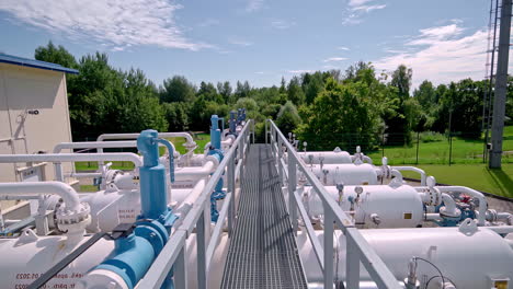 a walkway runs through rows of industrial gas or liquid storage tanks and pipelines, surrounded by greenery under a bright, clear sky - aerial pullback shot