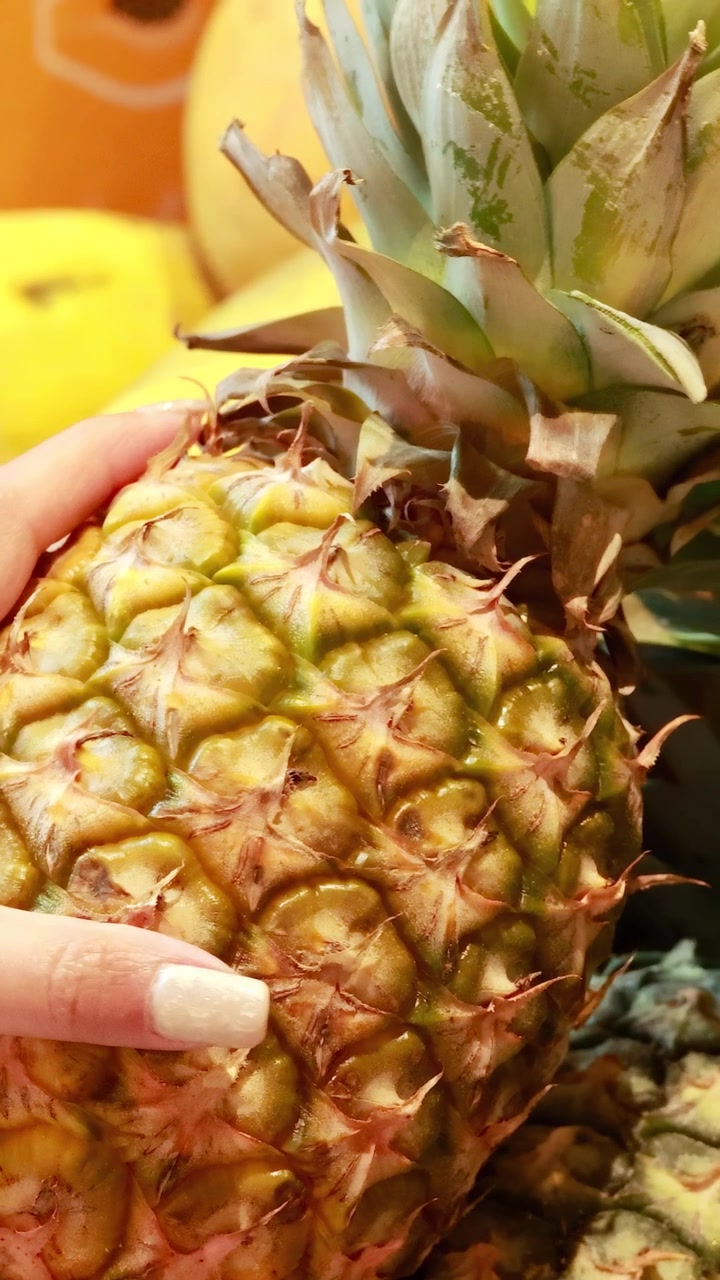 A person examines a pineapple in a brightly lit supermarket environment, focusing on texture and ripeness