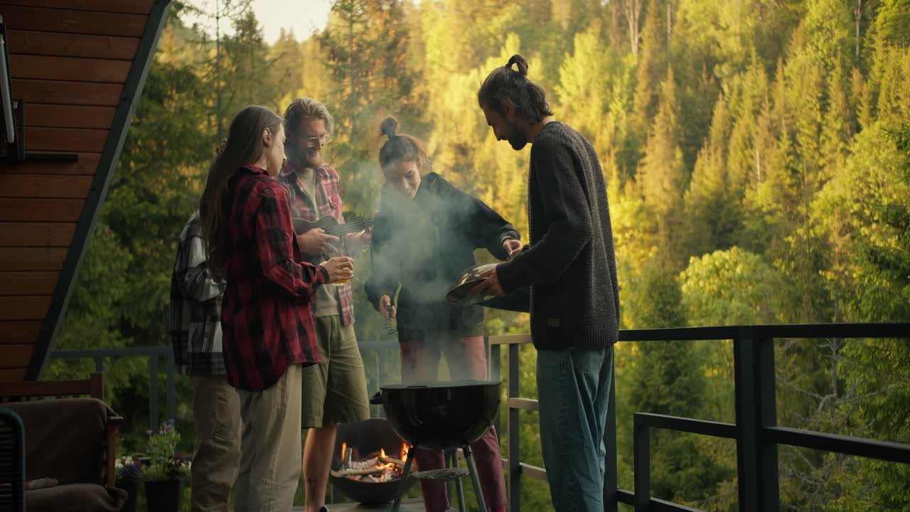 A group of friends are relaxing and grilling meat on the terrace of a country house against the backdrop of a green coniferous forest. The guy plays the guitar, a couple of people work with the barbecue