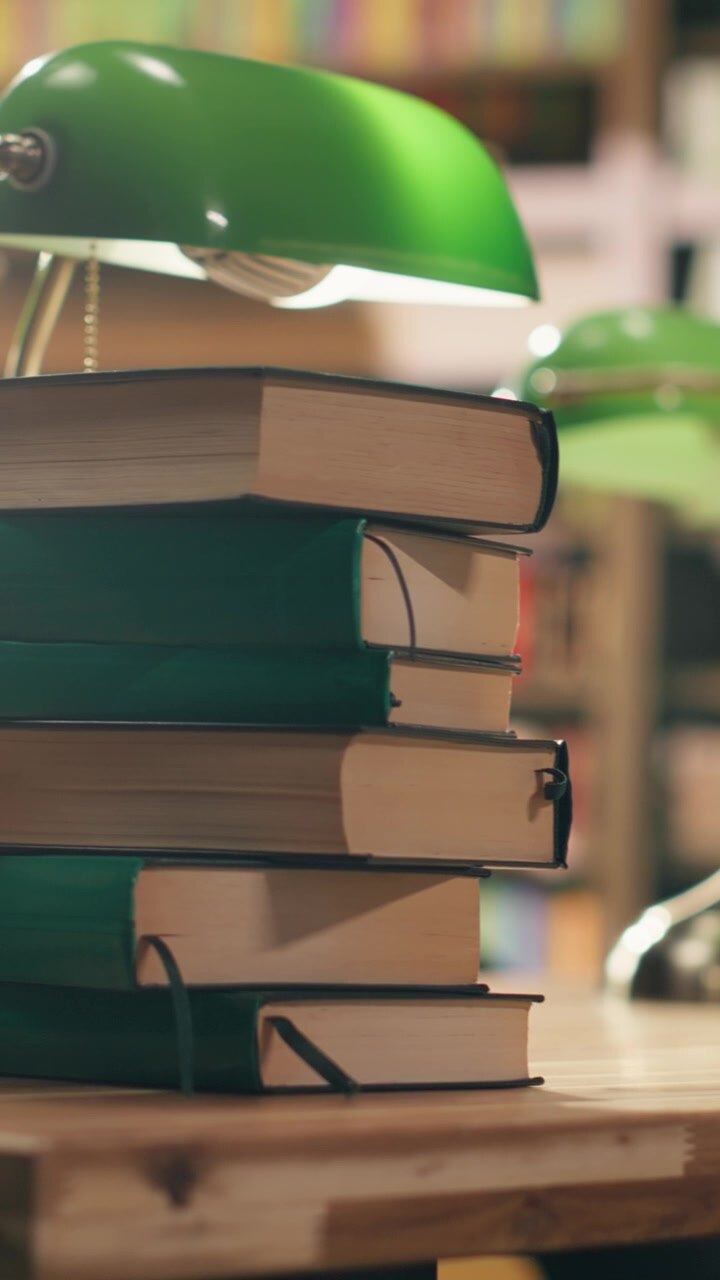 Video Vertical: Pila de libros sobre una mesa de biblioteca vacía con lámparas verdes cerca de las estanterías.