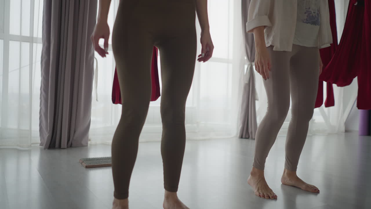 Downward view of yoga enthusiasts gracefully shifting legs and bodies in soft-lit studio with sheer curtains and red hammocks, emphasizing balance, rhythm, and fluid movement in mindful practice