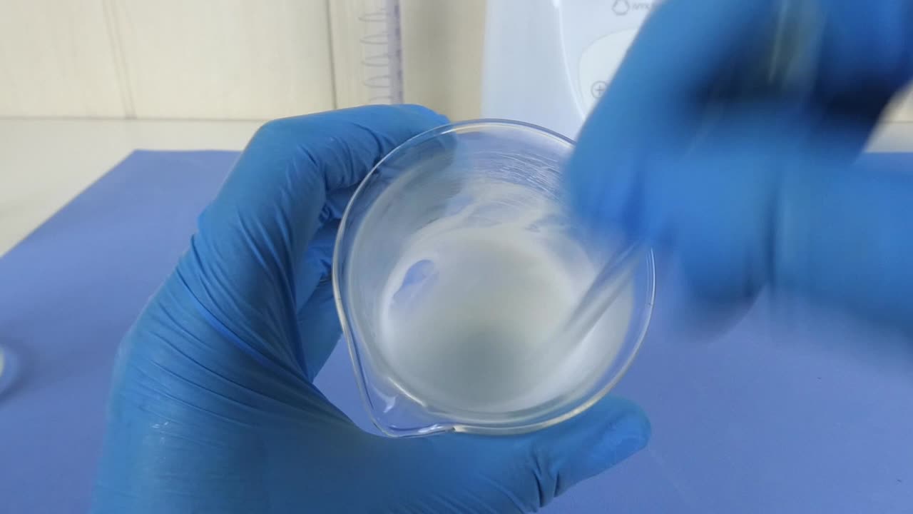 Macro shot of a cosmetic emulsion being prepared in a laboratory. The video captures the operator's gloved hands mixing ingredients in a beaker,