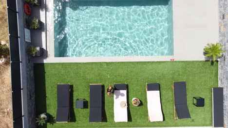 drone flies over an outdoor pool in bird's eye view - woman in swimming costume gets out of the pool and lies on sun lounger - villa in greece, crete with palm trees