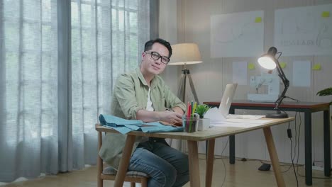 직물을 입은 아시아인 디자이너가 사무실에서 노트북을 사용하면서 레이아웃을 보고 팔을 고 카메라에 미소 짓고 있습니다.