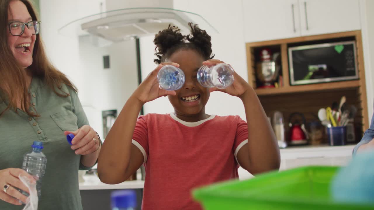 Happy caucasian lesbian couple and their african american daughter sorting waste in kitchen