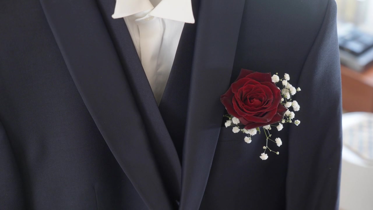 La telecamera si avvicina a una boutonnière di rosa rossa appuntata su un abito da sposo blu navy.