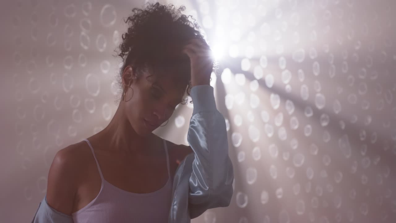 Une femme adulte se brossant les cheveux à contre-jour, faisant glisser sa veste de son épaule montrant un débardeur et des créoles en studio.
