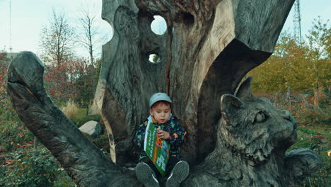 beautiful toddler boy sitting on a big wooden sculpture of cat. cute kid is focused on the colorful book.