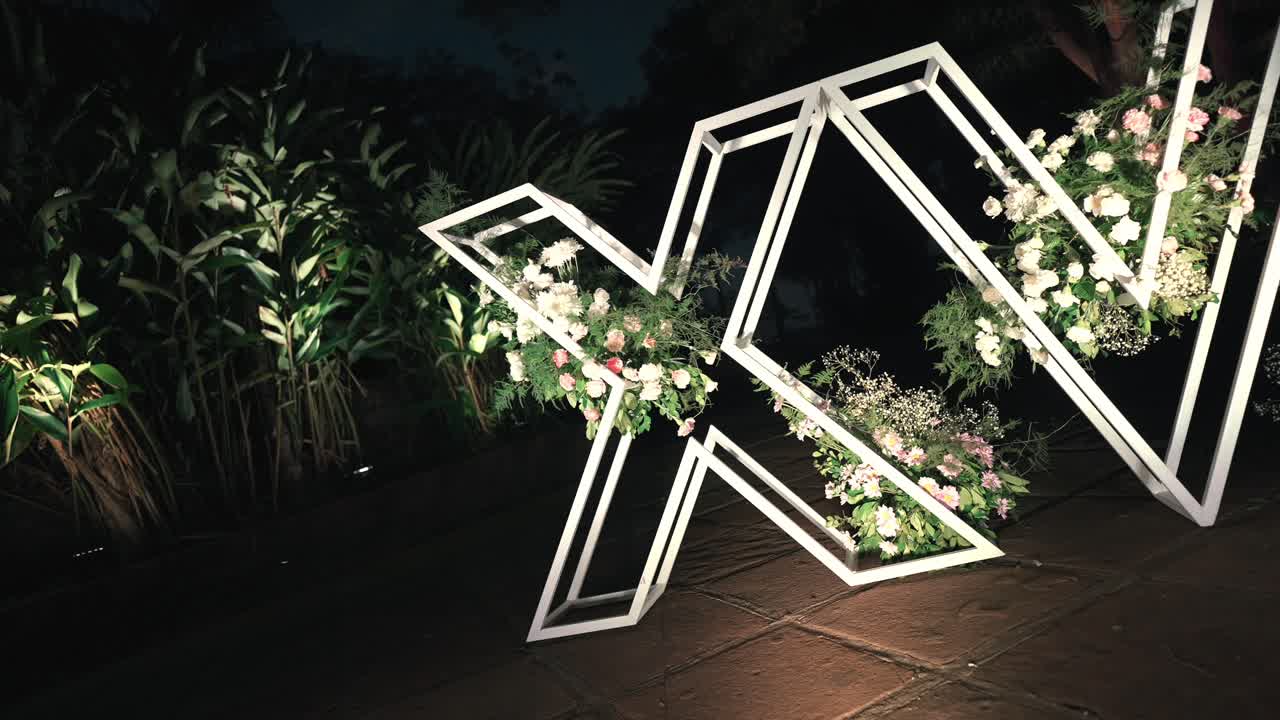 Angled camera shot of a "XV" (Sweet 15) white wire welcome structure decorated with flowers and illuminated by spotlights at an outdoor night party