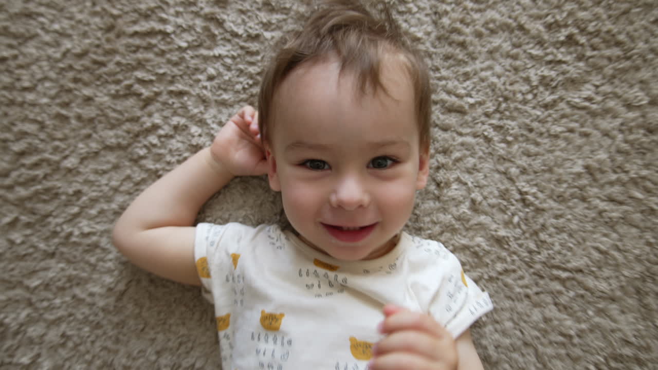 Camera approaches a beautiful Caucasian baby boy lying on the carpet. Kid smiles to camera and turns on right side. Top view.